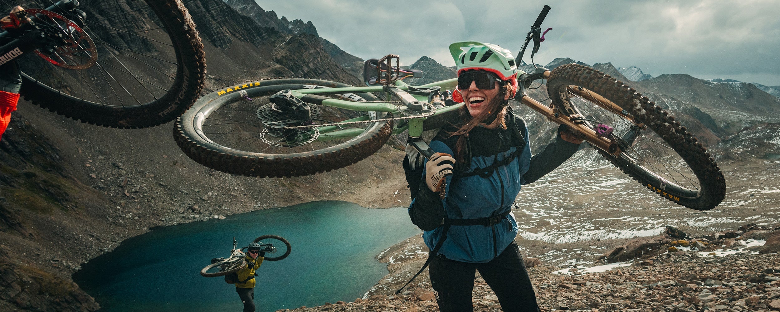 Female holding her bike on her back wearing Outdoor Research Women's Freewheel MTB Stretch Rain Jacket Galactic/Black and Freewheel MTB Leather Palm Bike Gloves Khaki. 