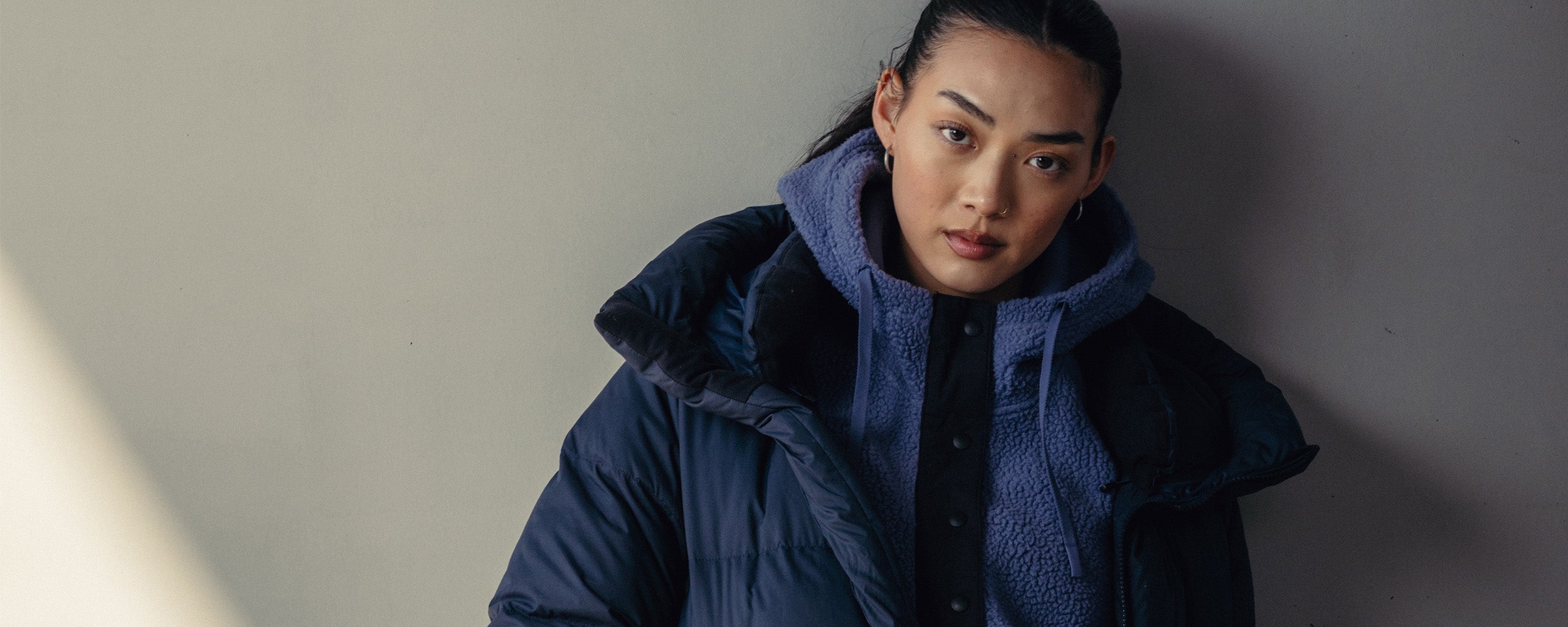 A woman stands by a wall wearing Outdoor Research Women's Coze Down Parka Atlantic and Women's Grayland Pullover Hoodie Summit. 