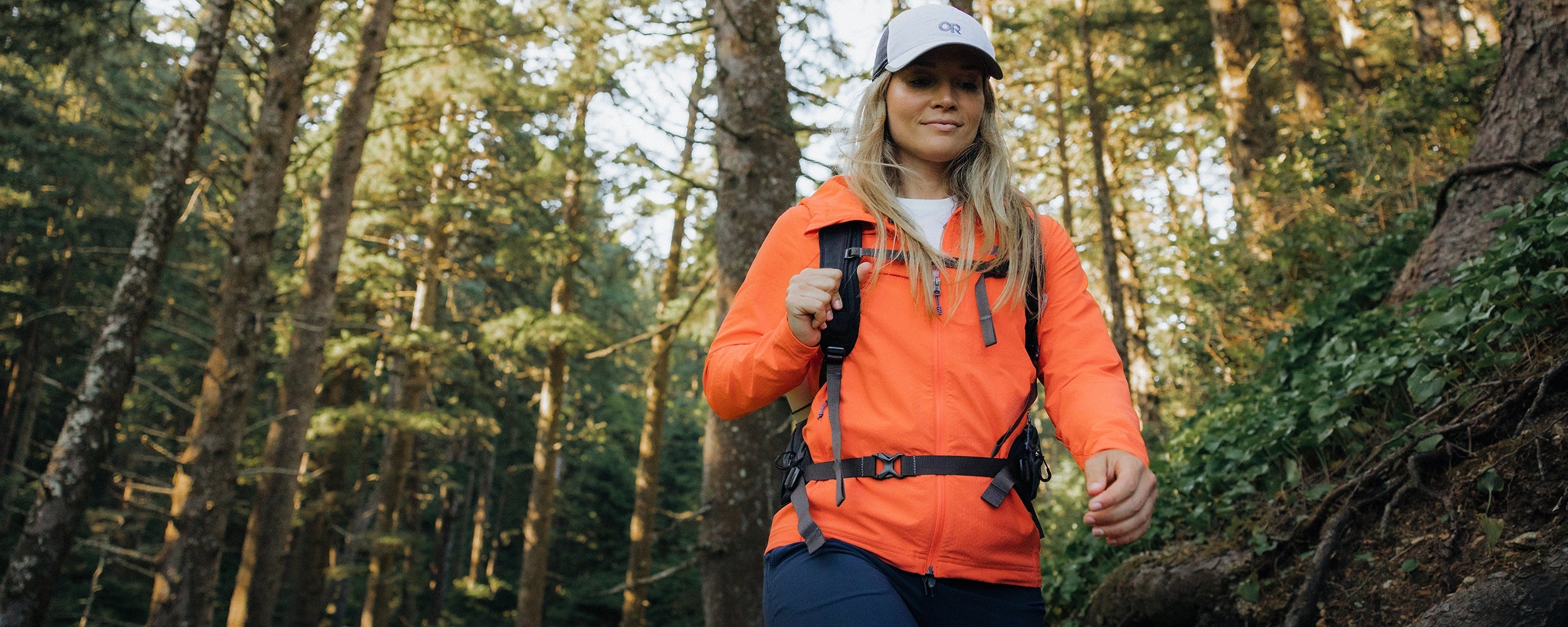 Woman hikes through the woods in the sun while wearing the Outdoor Research Ultima Softshell Hoodie in Zest/Orange. 