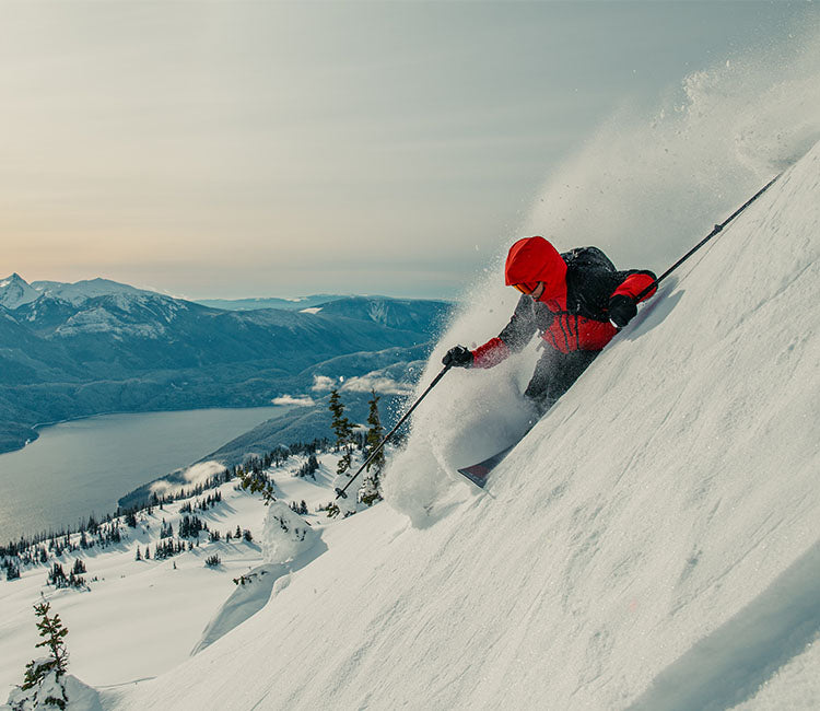 Skier going down steep mountain with a view of the water wearing Outdoor Research Men's Powderverse Jacket Lingonberry/Black. 
