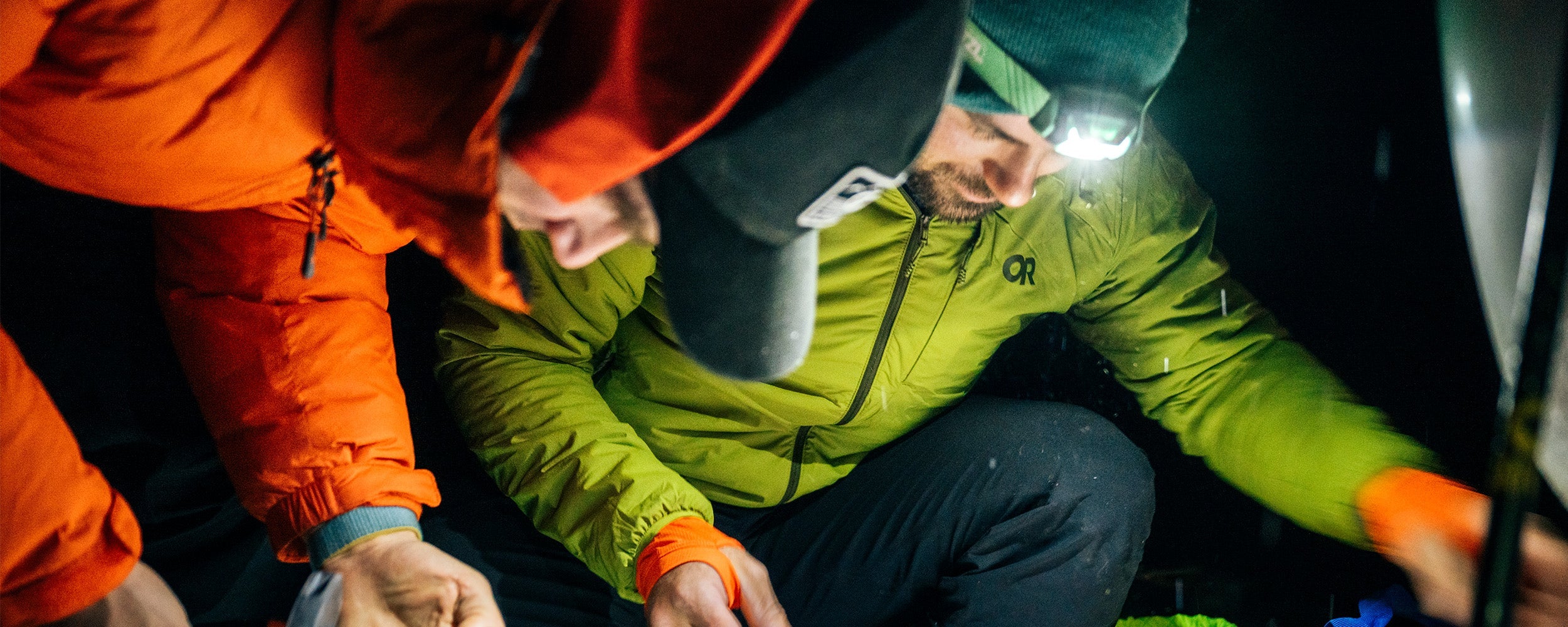 Two friends in the dark with flashlights looking down wearing Outdoor Research Men's Vesper Insulated Hoodie Rainforest Green. 