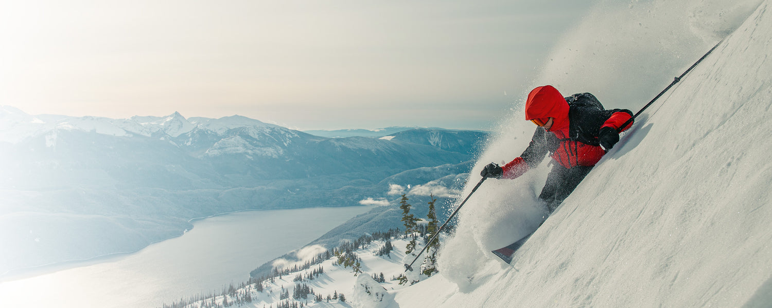 Skier going down steep mountain with a view of the water wearing Outdoor Research Men's Powderverse Jacket Lingonberry/Black. 