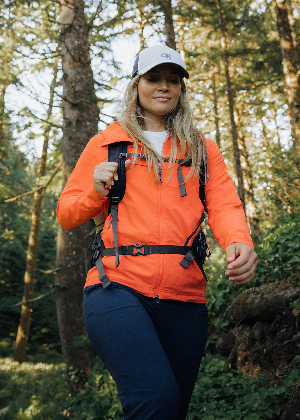 Woman hikes through the woods in the sun while wearing the Outdoor Research Ultima Softshell Hoodie in Zest/Orange. 